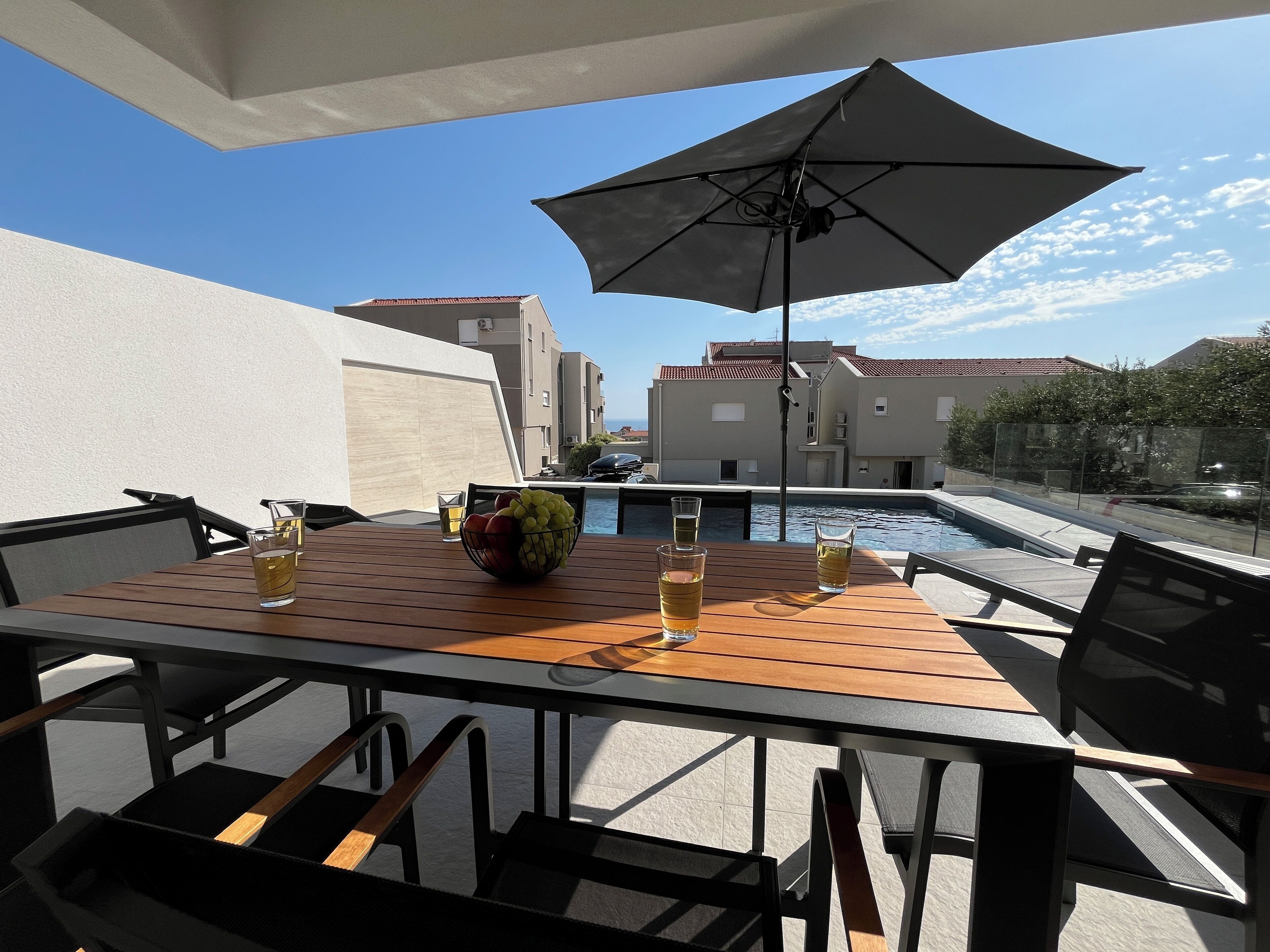 A wooden table with a bowl of fruit and glasses of beer, an umbrella, and a pool in the background.