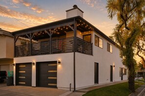 A modern two-story house with a balcony, garage doors, and string lights. - CDM Goldenrod - 5 min Walk to Beach with 2 Decks, Firepit, Central AC (Newport Beach)