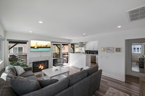 A modern living room with a fireplace, a large sofa, and a dining area with a view of the city. - CDM Goldenrod - 5 min Walk to Beach with 2 Decks, Firepit, Central AC (Newport Beach)