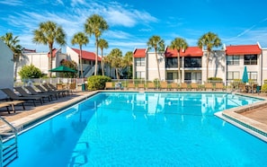 A swimming pool with lounge chairs and umbrellas, surrounded by palm trees and buildings with red roofs. - Pickleball • Private Beach Access • Largest Pool (Bradenton Beach)