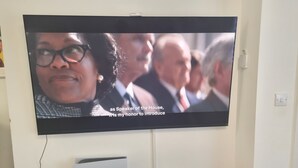 Un téléviseur fixé au mur affiche une scène où l’on voit des personnes.