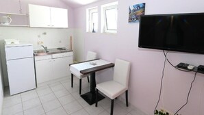 Une cuisine compacte avec des meubles blancs et un évier, une table à manger avec deux chaises, un téléviseur à écran plat fixé au mur et une petite plante en pot.