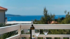 A balcony with a bottle of wine and two glasses, overlooking a coastal view with a clear sky.