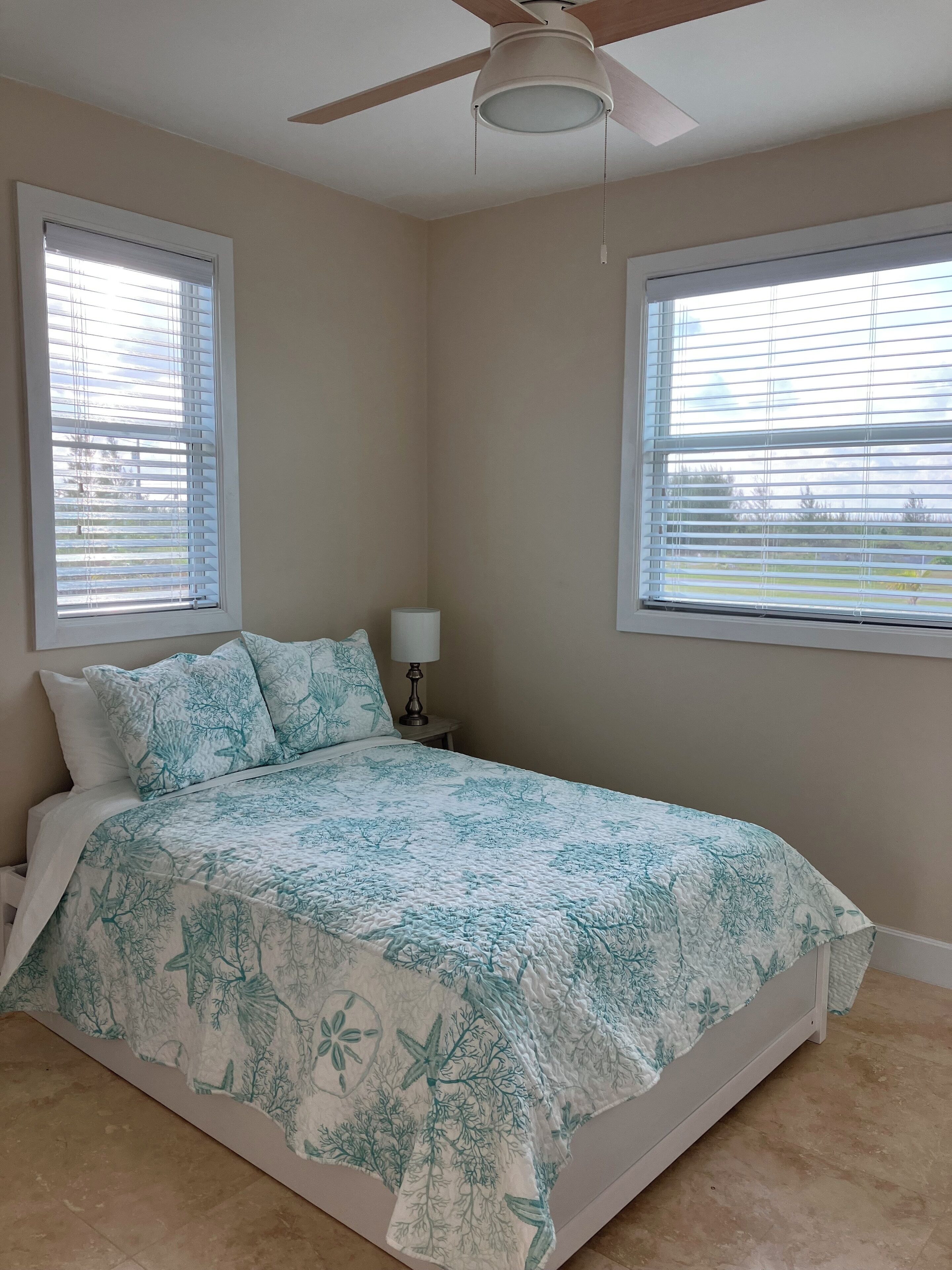 Quarto com cama, duas janelas com persianas, ventilador de teto e mesa de cabeceira com abajur.