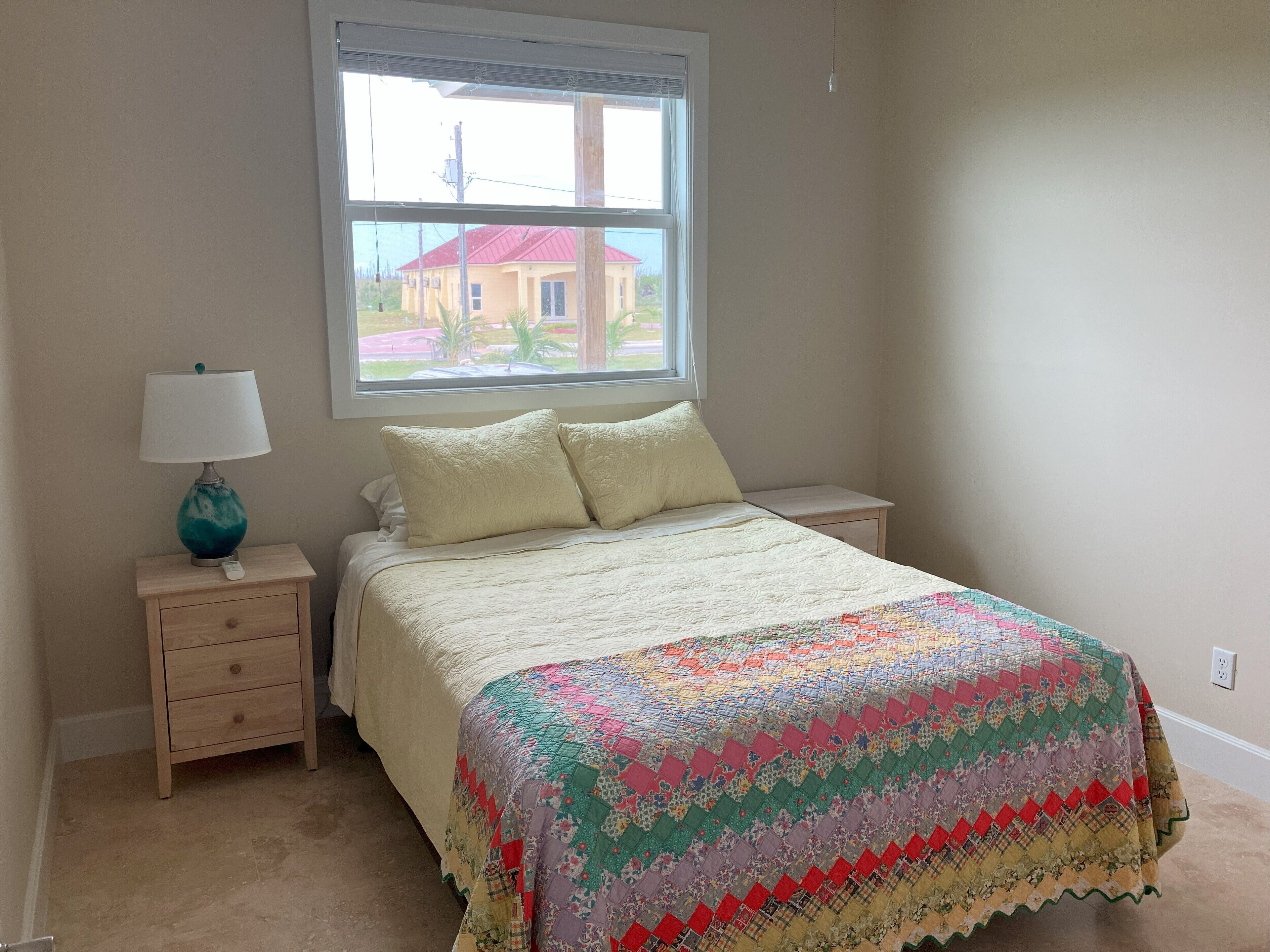 A bedroom with a bed, a nightstand, a lamp, and a window with a view of a house.