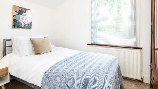 A bedroom with a bed, a nightstand, a radiator, a window with blinds, and a door. - Hanwell Private Single Room Near Station by Omega (London)