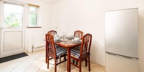 A dining area with a wooden table set for four, surrounded by chairs with striped cushions. A white refrigerator is to the right, and a window with blinds is on the left.