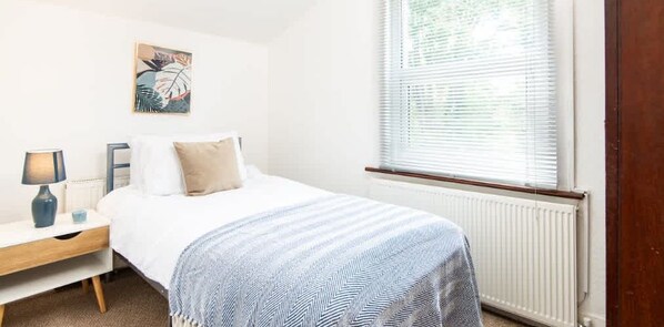 A bedroom with a bed, a bedside table, a lamp, a radiator, and a window with blinds.