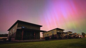 A modern house with a flat roof and a large porch area, set against a night sky with visible stars and a colorful aurora display.