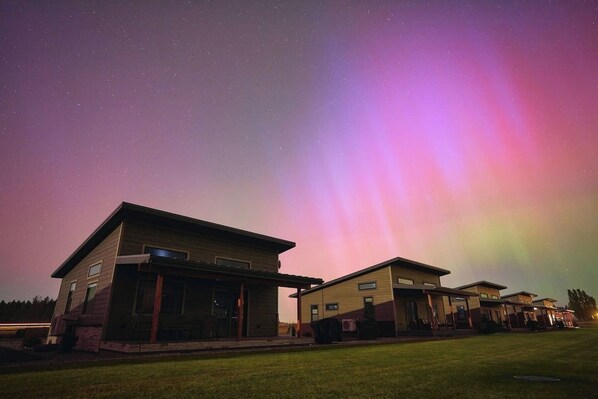 A modern house with a flat roof and a large porch area, set against a night sky with visible stars and a colorful aurora display.