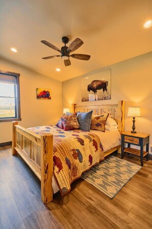 A bedroom with a wooden bed frame, a ceiling fan, a mounted artwork of a buffalo, and a nightstand with a lamp.