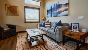 A living room with a sofa, a coffee table, a rug, a map on the wall, and a window with a view of the sky.