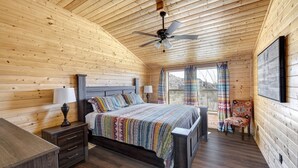 Een slaapkamer in een houten blokhut, met een bed, nachtkastjes, een stoel, een plafondventilator en een raam met gordijnen.