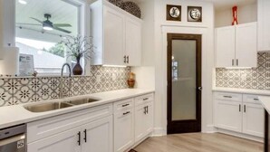 A kitchen with white cabinets, a patterned backsplash, and a window with a ceiling fan. - Gulf Breeze Retreat#Beaches#vacation rental. (Gulf Breeze)
