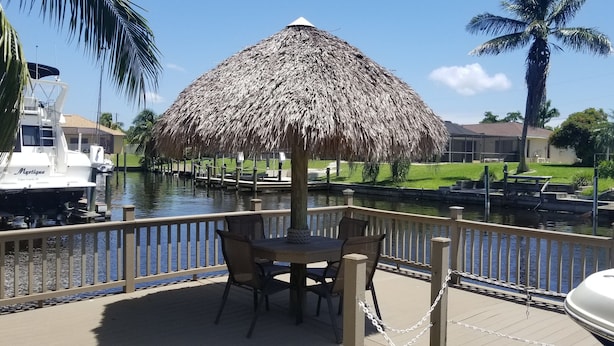 A boat docked by a canal, a thatched umbrella, and a table with chairs on a wooden deck.