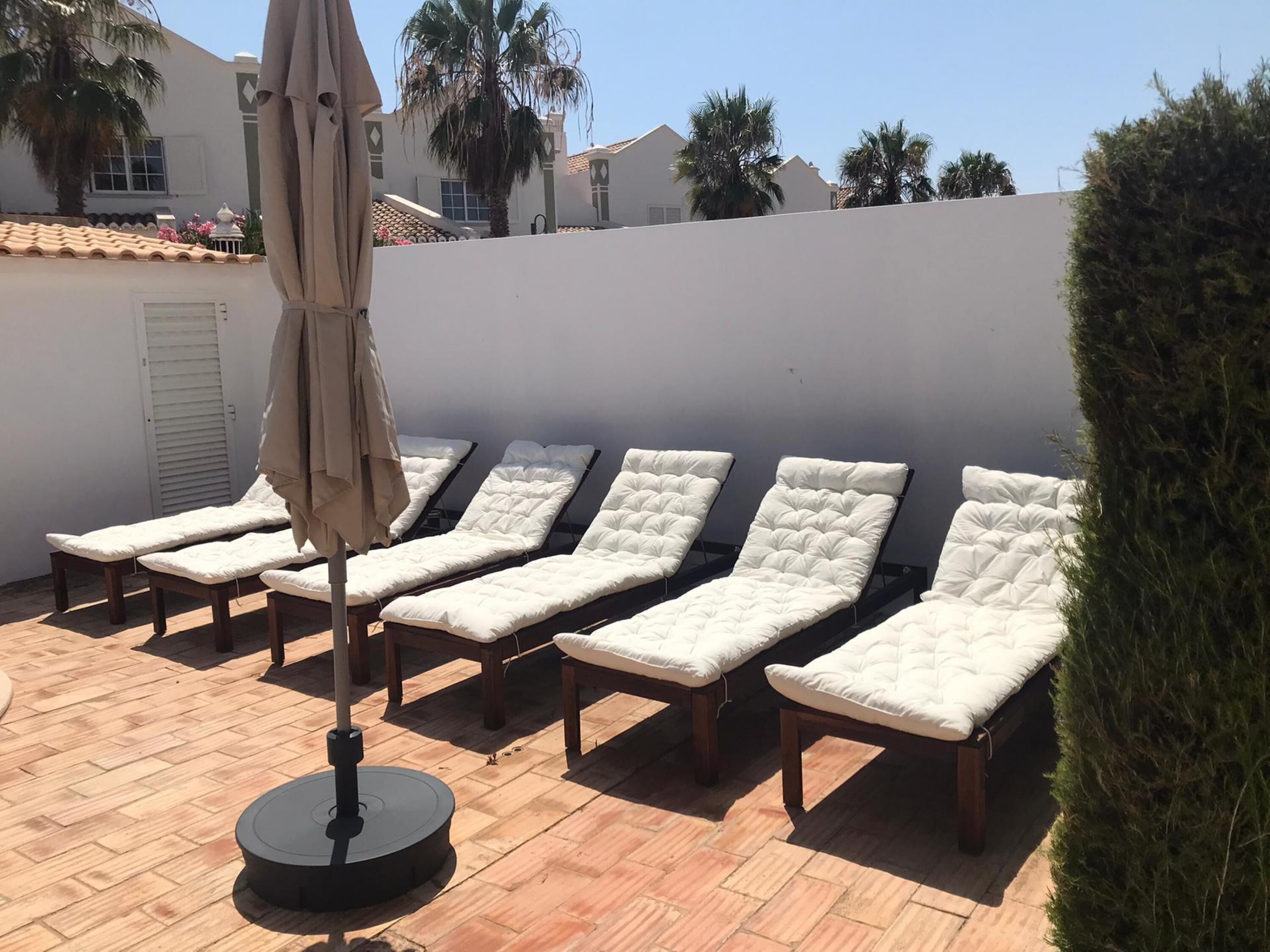A row of six sun loungers with cushions, a round table, and an umbrella on a tiled patio.