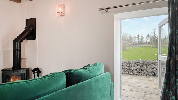 Ein gemütliches Wohnzimmer mit einem grünen Sofa, einem Kaminofen und Blick auf eine steile Wand und ein saftiges Grasfeld durch eine Glastür.