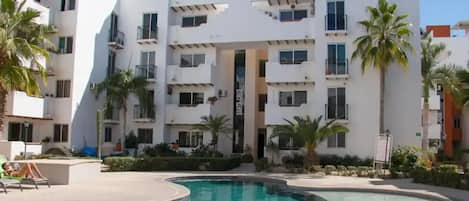 A modern apartment building with a swimming pool, balconies, and palm trees.