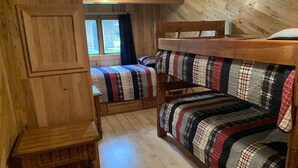 A cabin-style room with wooden floors, a bunk bed with plaid bedding, a small wooden table, and a window with curtains.