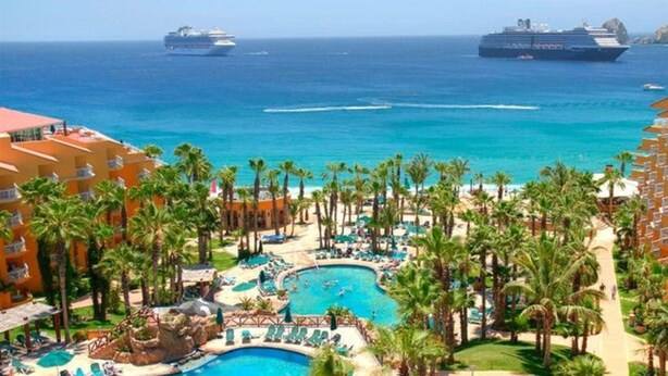 Een resort met meerdere zwembaden, palmbomen en uitzicht op de oceaan met cruiseschepen.