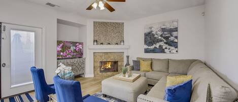 A modern living room with a fireplace, a ceiling fan, and a large window.