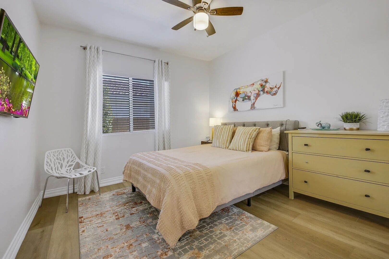 A bedroom with a bed, a dresser, a chair, a ceiling fan, a window with blinds, and a painting on the wall.