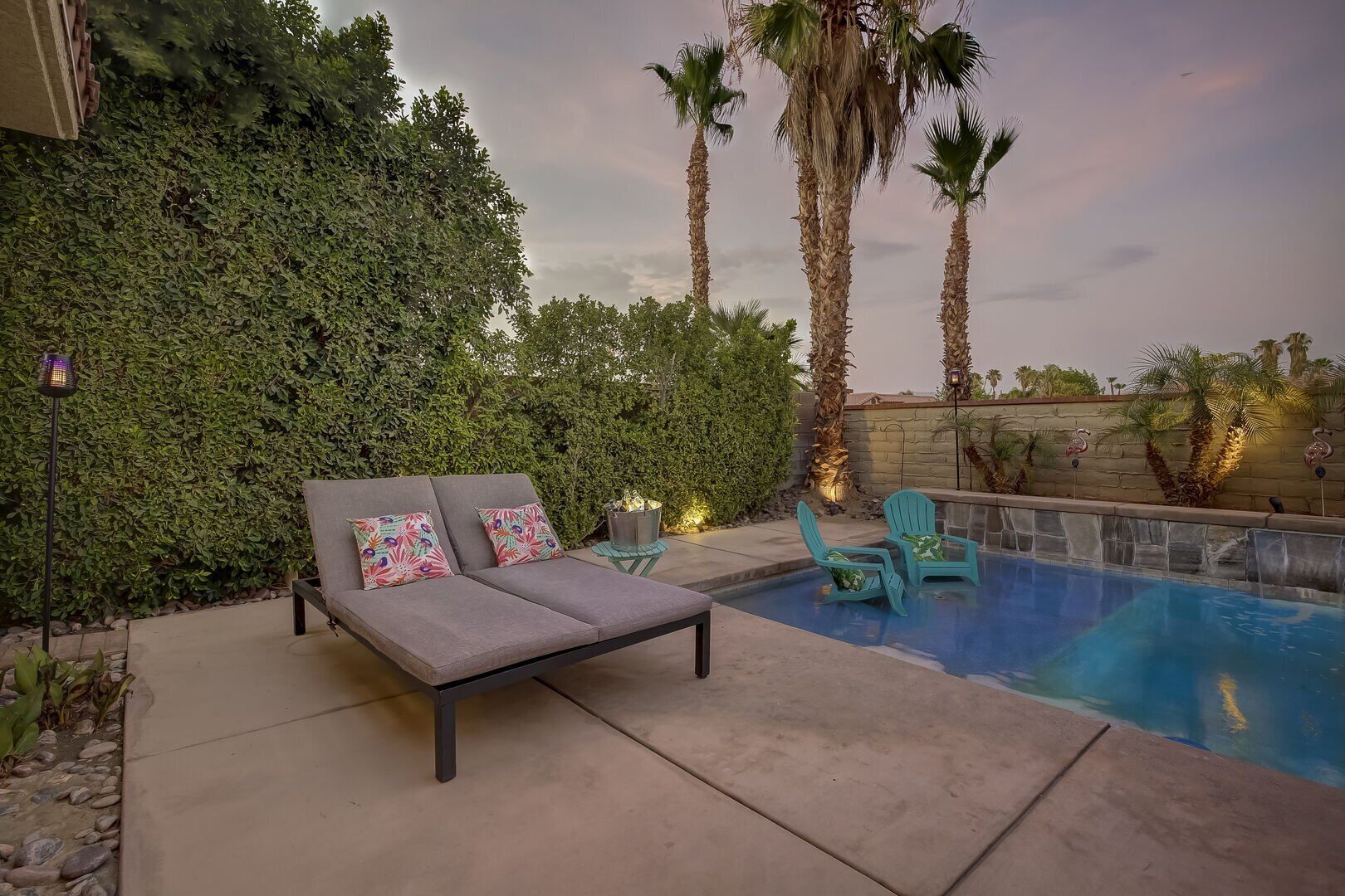 A pool area with a lounge chair and two chairs, surrounded by palm trees and a hedge.