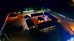 Vista aérea nocturna de un complejo de edificios con varias estructuras, incluyendo un edificio central con interior iluminado, rodeado de edificios más pequeños y un área cubierta.