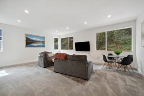 Ein modernes Wohnzimmer mit einem grauen Sofa, einem Flachbildfernseher und einem Essbereich mit Tisch und Stühlen.