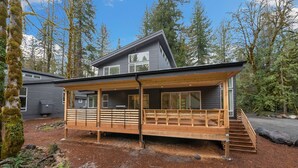 Una cabaña moderna con terraza de madera, ventanas amplias y techo pronunciado, rodeada de bosque denso.