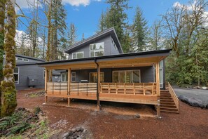 A modern cabin with a wooden deck, large windows, and a steeply sloped roof, surrounded by dense forest. - Sandy River Group Getaway 2 (Mount Hood Village)