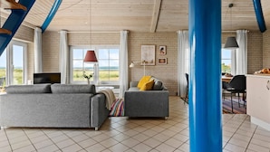 Ein großzügiger Wohnbereich mit einem großen Fenster und Blick auf den Strand, ein graues Sofa, ein kleinerer grauer Sessel und ein Essbereich mit Tisch und Stühlen.