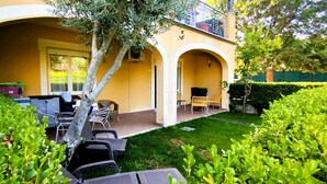 Een geel gebouw met een overdekte patio, zitplaatsen buiten en een verzorgde tuin.
