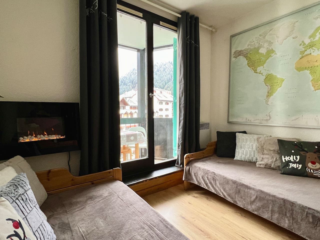 Una habitación acogedora con dos camas individuales, piso de madera, chimenea, un ventanal grande con cortinas y un mapa del mundo en la pared.