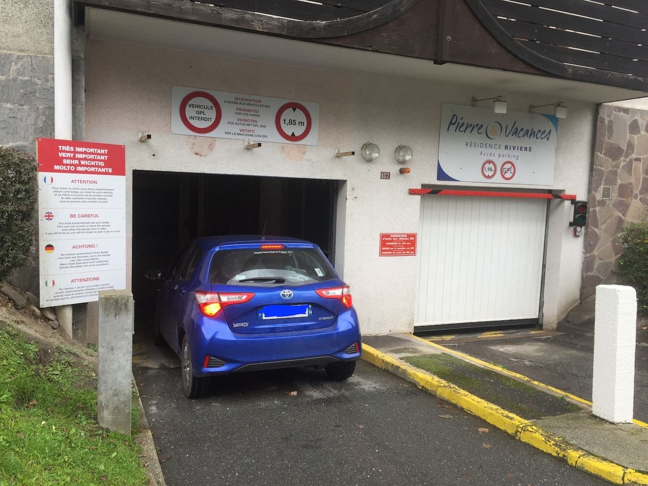 Un coche azul entrando a un edificio con señales que indican un límite de altura de 1,85 metros y un área de estacionamiento para vehículos con GIPL.