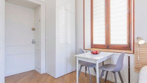 Een kleine, lichte kamer met een witte deur, een witte tafel met twee stoelen, een raam met zonwering en een lamp op een nachtkastje.