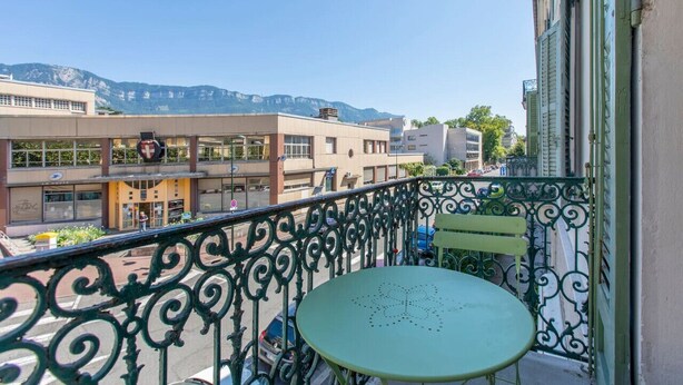 Een balkon met een groene tafel en stoel, met uitzicht op een straat met gebouwen en bergen op de achtergrond.