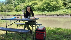 Um piquenique às margens de um rio, com mesa, banco e uma sacola vermelha.
