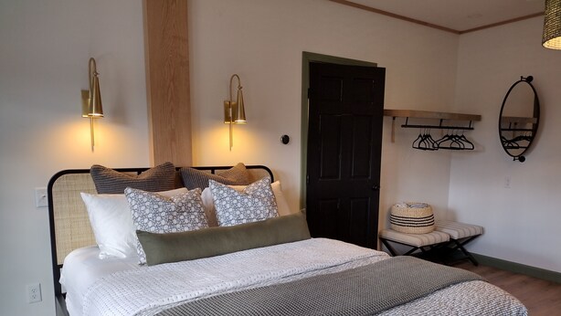 Uma cama bem arrumada com colcha estampada, duas luminárias fixadas na parede, uma porta escura, um pequeno banco e uma cesta de palha.
