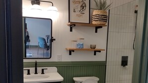 Casa de banho com uma sanita branca, lavatório, espelho e zona de duche. Existem prateleiras sobre o lavatório com objetos decorativos.