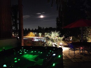 Un jacuzzi iluminado con luces verdes, un patio con un paraguas rojo y vistas a un estacionamiento con árboles y la luna en el cielo.