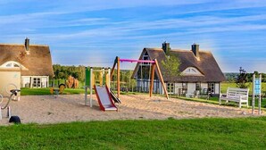 A playground with a swing set and slide, surrounded by houses with thatched roofs and a grassy area.
