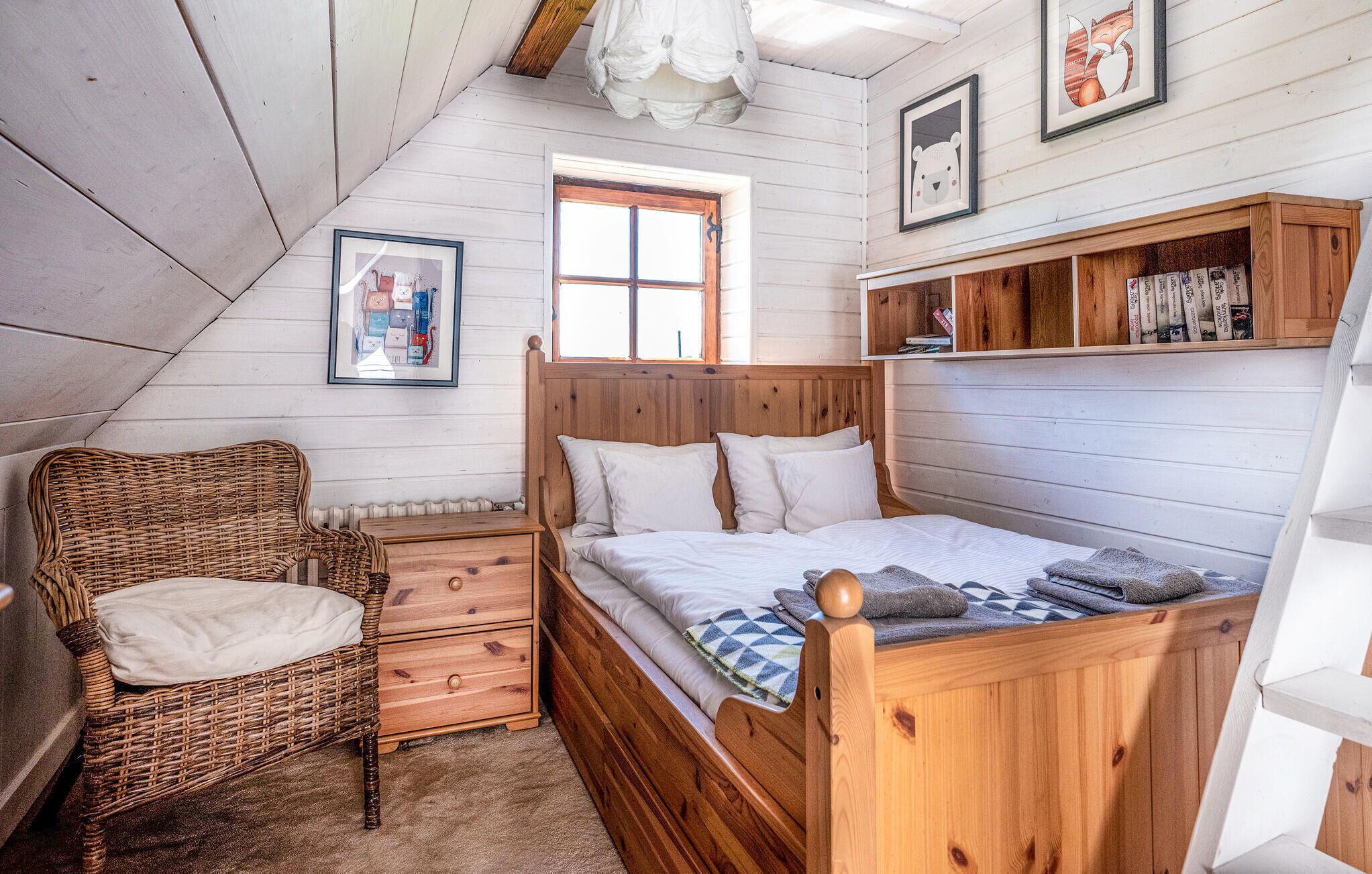 A cozy bedroom with a wooden bed, a wicker chair, a nightstand, and a shelf with books.