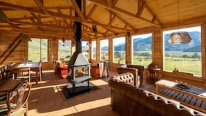 A wooden cabin interior with a fireplace, large windows, and a view of mountains. - Estancia Bonanza Patagonian Lodge Experience (El Chalten)