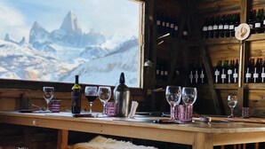 A wooden bar with wine glasses and bottles, a view of snow-capped mountains, and a wooden shelf with wine bottles. - Estancia Bonanza Patagonian Lodge Experience (El Chalten)