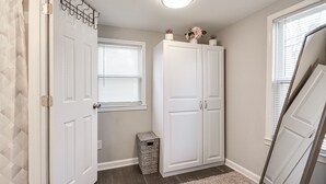 Un couloir avec une porte blanche, une fenêtre avec des stores, une armoire blanche et une petite poubelle.