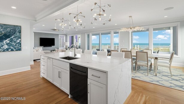 Una cocina moderna con una isla grande, armarios blancos y un lavavajillas integrado. Hay un comedor con mesa y sillas, y un ventanal con vistas al océano.
