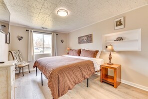 Ein Schlafzimmer mit einem großen Bett, einem Nachttisch mit Lampe, einem Sessel, einem Schreibtisch und einem Fenster mit Vorhängen.