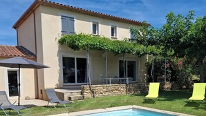 A swimming pool with a stone border, a patio area with chairs, and a house with a tiled roof and windows.