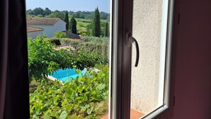 A window with a view of a swimming pool surrounded by greenery and a distant landscape.
